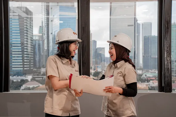 Dos ingenieras con casco revisando planos técnicos en una oficina.